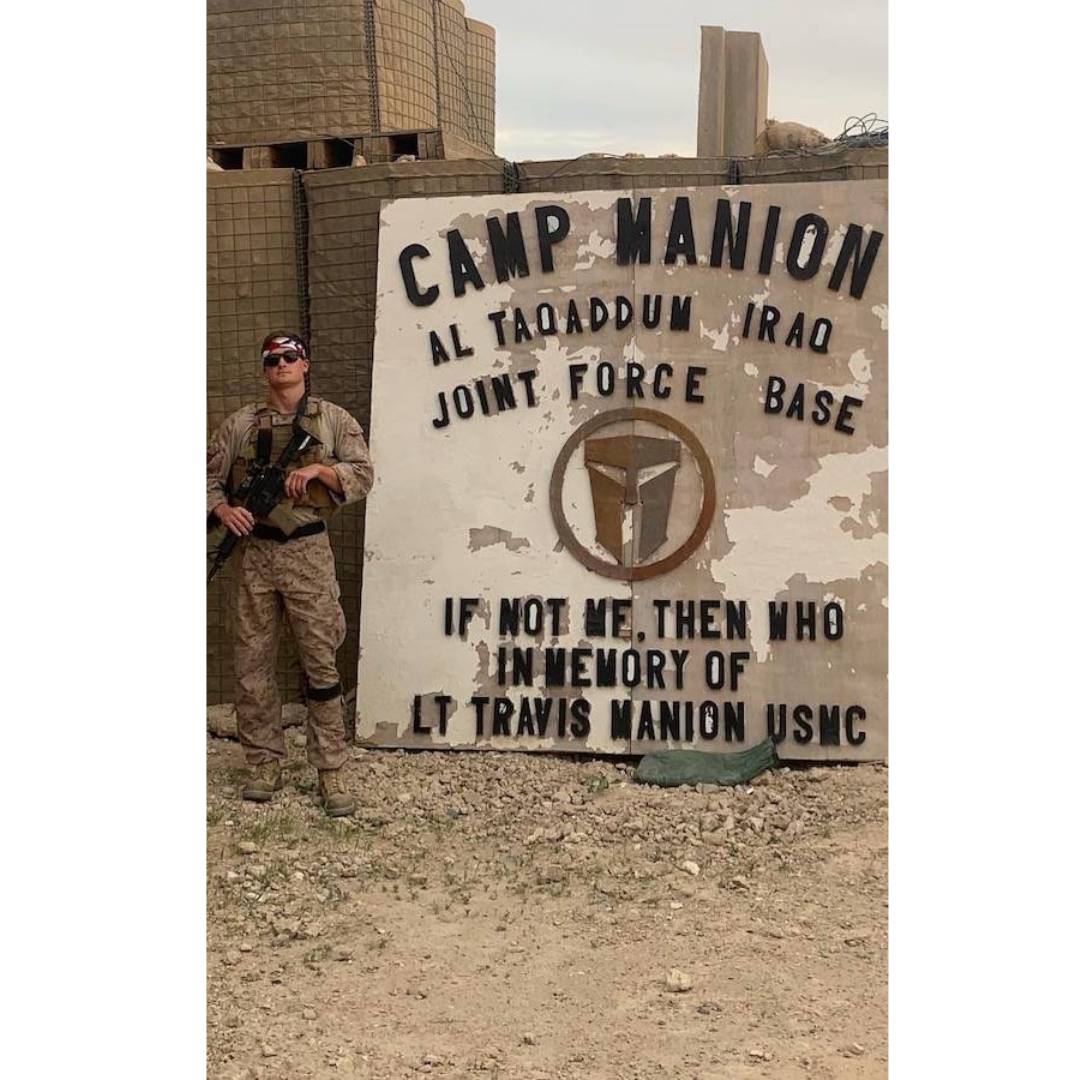 Cameron Umphrey standing in front of a sign that says Camp Manion Al Taqadum Iraq Joint Force Base If Not Me, Then Who In Memory of LT Travis Manion USNC and he is standing near a flag